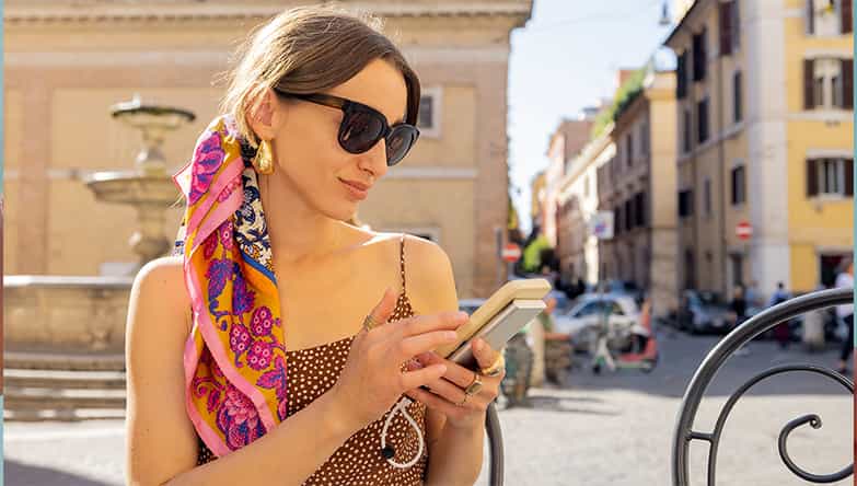 Une femme portant des lunettes de soleil est assise sur une terrasse en utilisant son téléphone et une batterie externe.