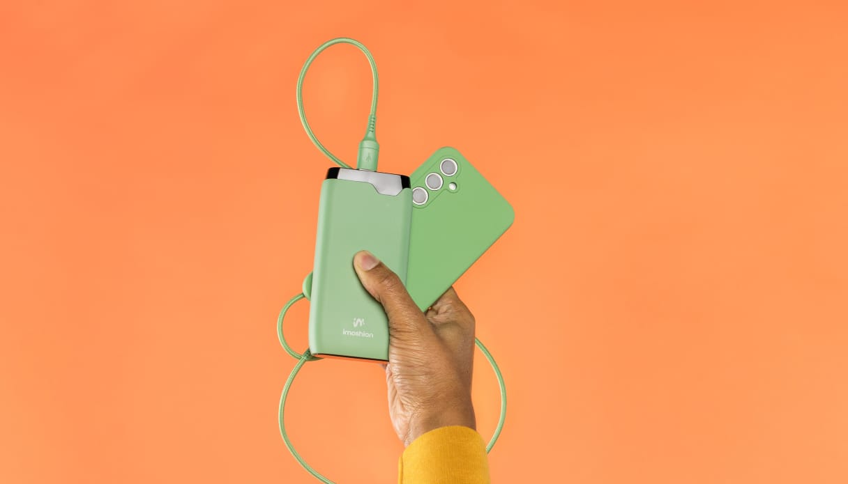 Une main dont on aper&ccedil;oit un bout de manche jaune sort du bas de l'image et tient un t&eacute;l&eacute;phone avec une coque verte, un c&acirc;ble de recharge vert et une batterie externe imoshion verte. L'arri&egrave;re-plan est orange.