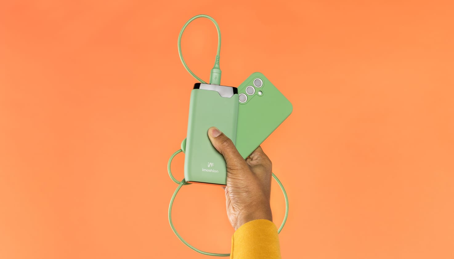 Une main dont on aper&ccedil;oit un bout de manche jaune sort du bas de l'image et tient un t&eacute;l&eacute;phone avec une coque verte, un c&acirc;ble de recharge vert et une batterie externe imoshion verte. L'arri&egrave;re-plan est orange.