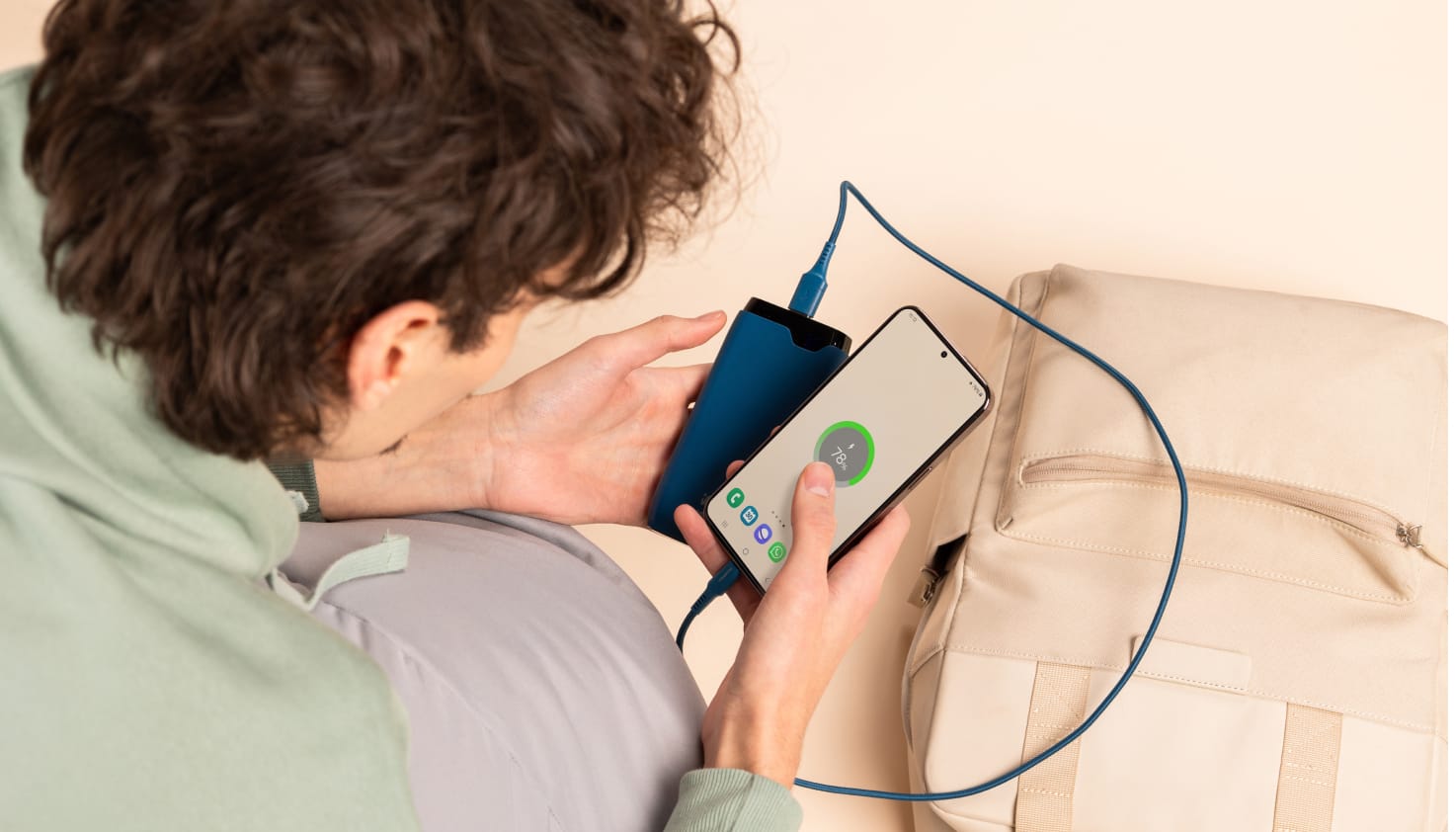 Vous voyez une personne aux cheveux bruns regardant un smartphone qui se recharge via un c&acirc;ble bleu branch&eacute; sur une batterie externe bleue. Sur le fond beige se trouve un sac &agrave; dos couleur sable.