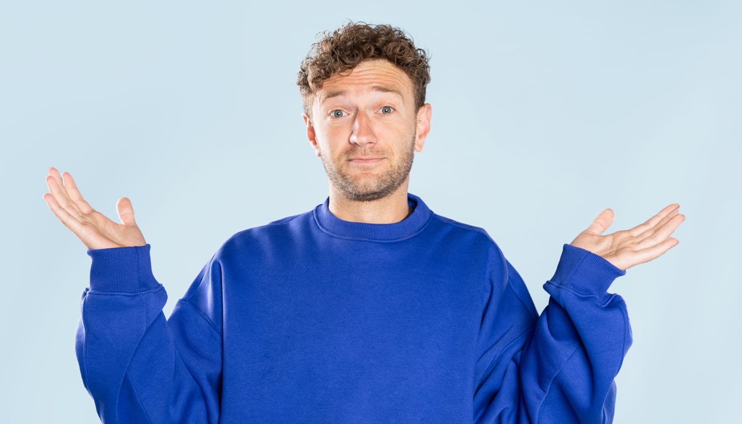 Un homme vêtu d'un pull bleu cobalt lève les mains en signe d'interrogation.